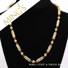 Ming's gold necklace with alternating cylindrical and round pierced beads on a black bust.